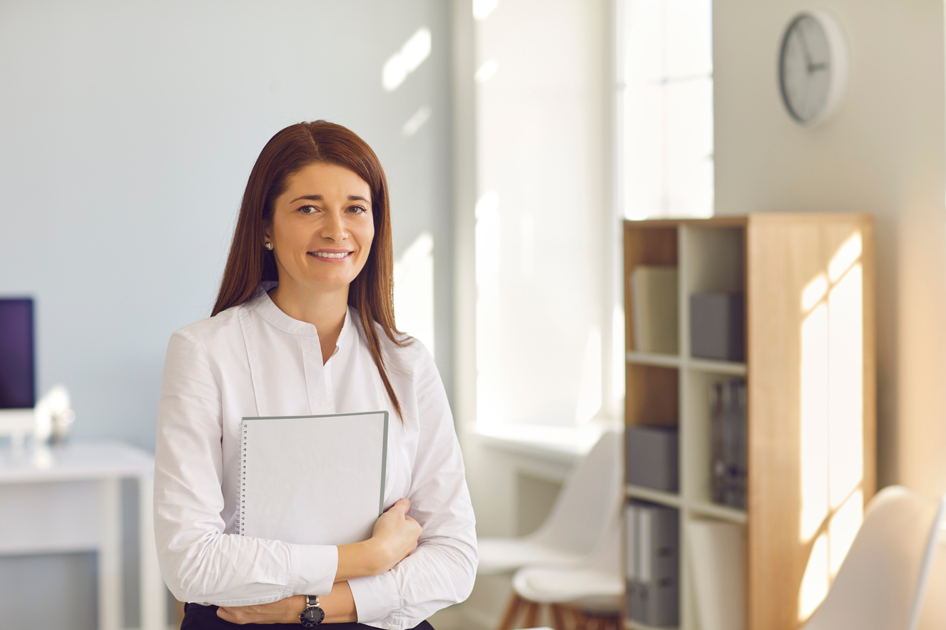 Smiling Business Woman with Notepad in Hands Looking at Camera Standing in Sunny Company Office